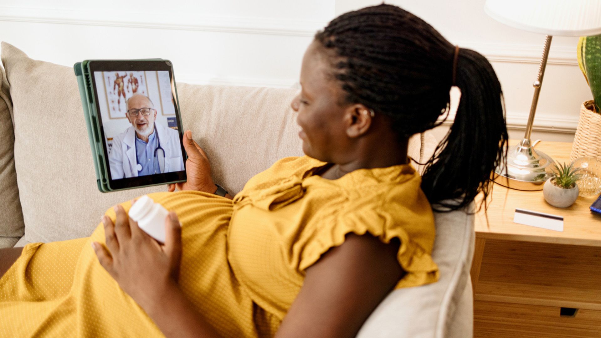 — title missing — Pregnant woman sitting on a couch with a bottle of medicine talking with her doctor via a tablet.