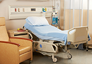 iStock-hospital-bed-180px.jpg Empty hospital bed in a room with a chair, table, and medical equipment next to it.