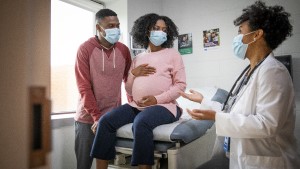 Couple_Expecting_Doctor_Masks Man with pregnant woman wearing masks and talking with a masked doctor in exam room