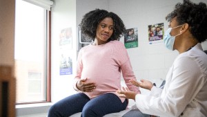 Pregnant_Woman_Doctor_Mask Pregnant Black woman sitting in exam room talking with doctor who is weaking a mask