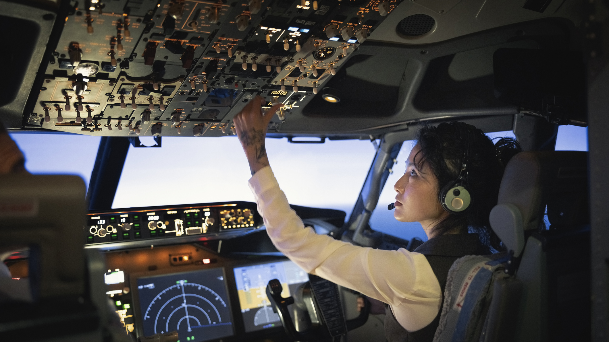 1432221653 Rear view of a female pilot adjusting switches on the control panel while sitting inside cockpit.