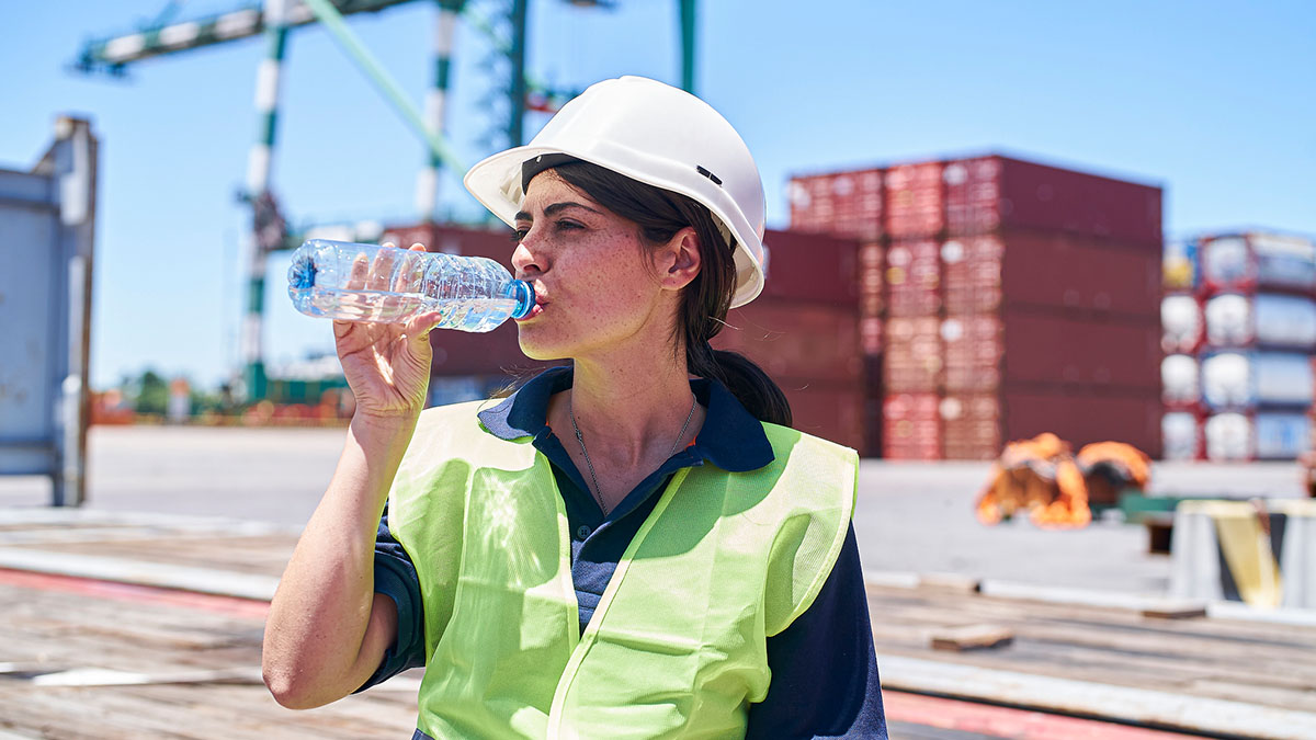 Workplace Recommendations Female construction wearing a white hard hat and yellow safety vest, drinking water while working outside