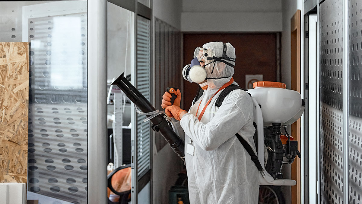 Man wearing a white protective suit disinfecting a work space