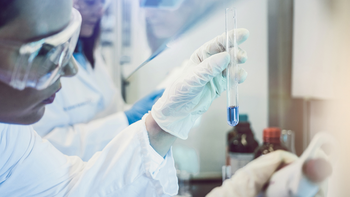 579776364 Scientist wearing gloved and eye protection, looking at a test tube.