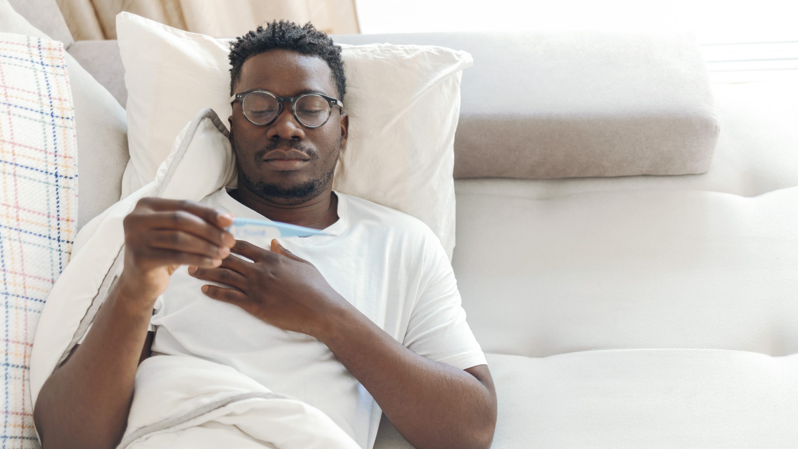 — title missing — African American man laying on a white couch, taking his temperature.