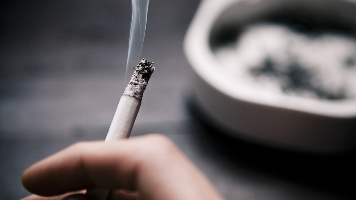 — title missing — a hand holding a cigarette in the foreground, and a blurry ashtray in the background.