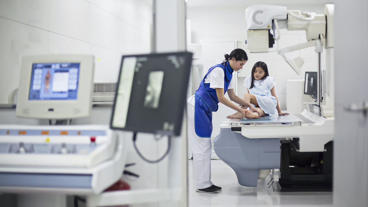 — title missing — A radiology technologist wearing protective lead drapes positions a child for an x-ray