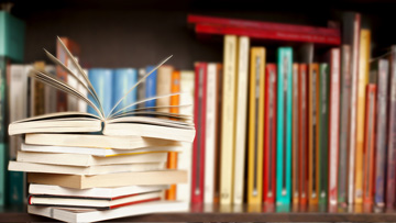 — title missing — Stack of books on a wooden library shelf, one of them open on top, multicolored book spines background.