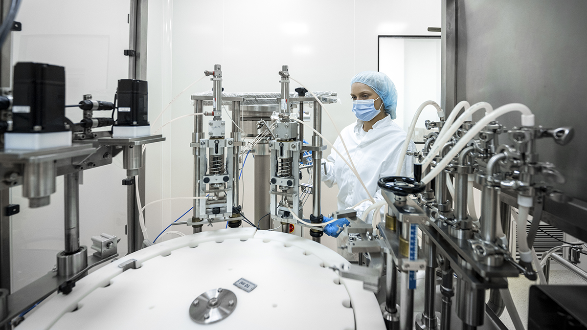 — title missing — Small brown bottles seen in a laboratory machine during manufacturing in a pharmaceutical factory while a female employee in a protective clothing is controlling the process.