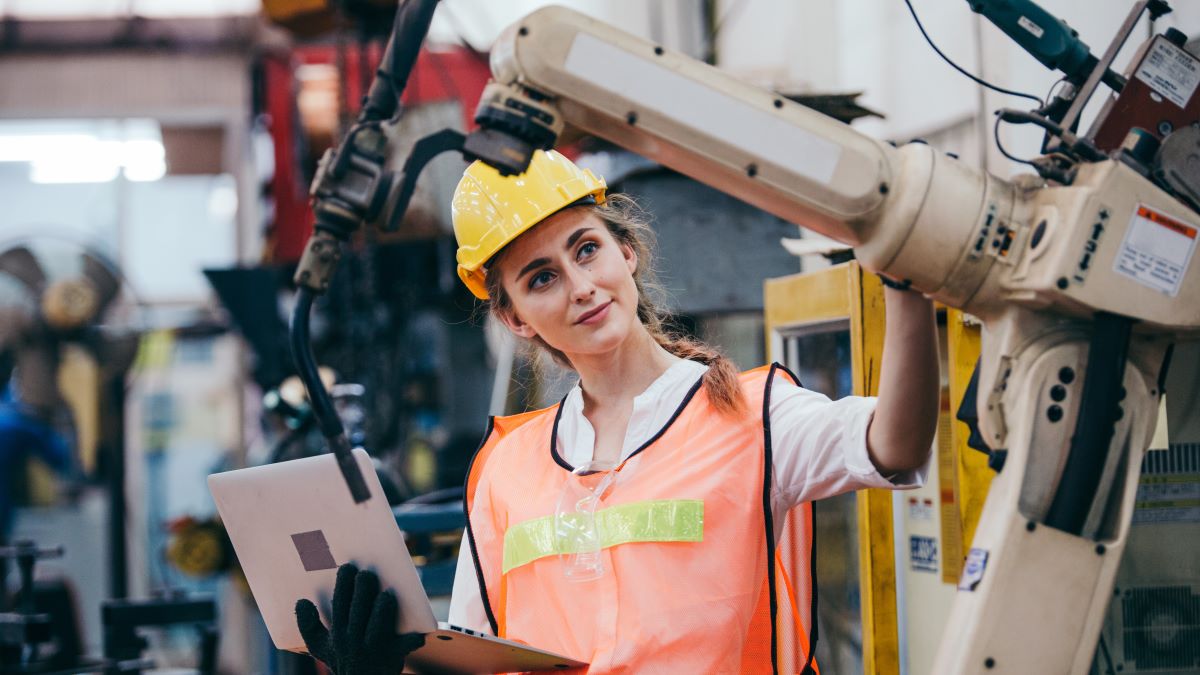— title missing — Woman wearing a hard hat and safety vest working next to a robotic arm.
