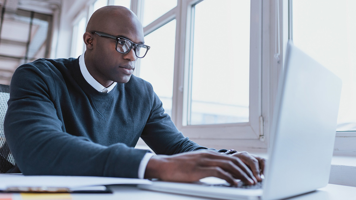 computer-img Man wearing glasses and a sweater, working on a laptop at a desk near a window.