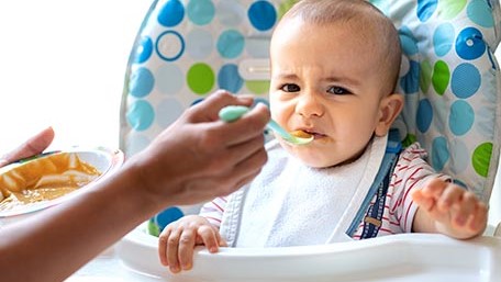 — title missing — Child in high chair being fed by an adult.