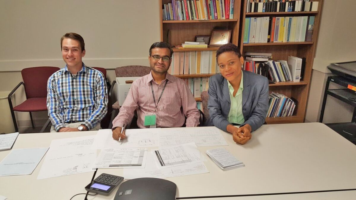 — title missing — Three people sitting a large table strewn with papers in an office setting with bookshelves.