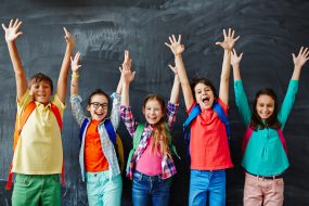 Happy Children Five smiling students in front of a chalkboard stretching their arms over their heads.