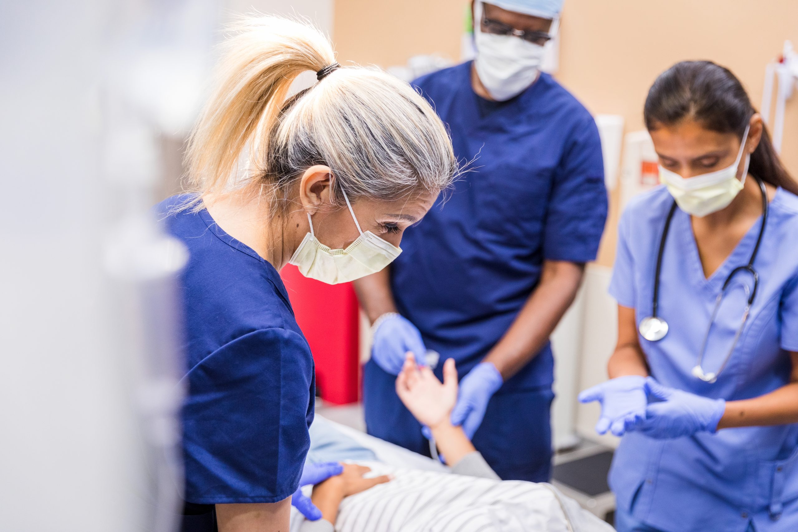 A three-person healthcare team stands around the bed of a patient as they administer care