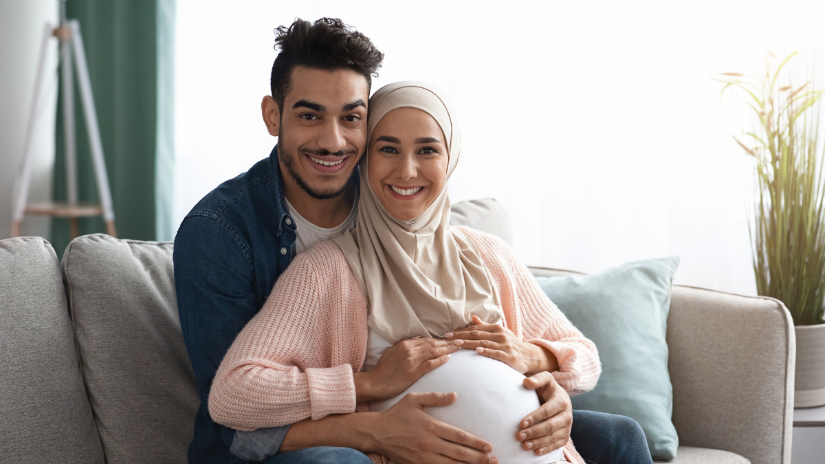 Pregnant couple Pregnant woman wearing hijab sitting on couch with man embracing her from behind with hands on her belly.