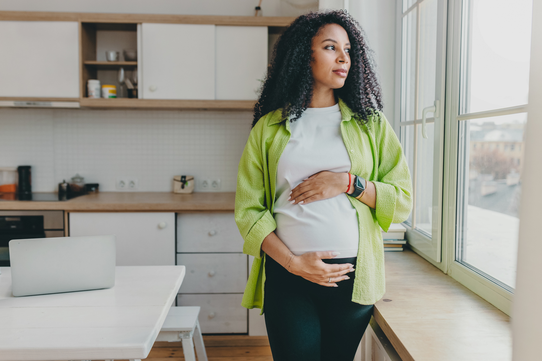 pregnant mother by window Pregnant woman of color standing and looking out the kitchen window, while holdingbelly.