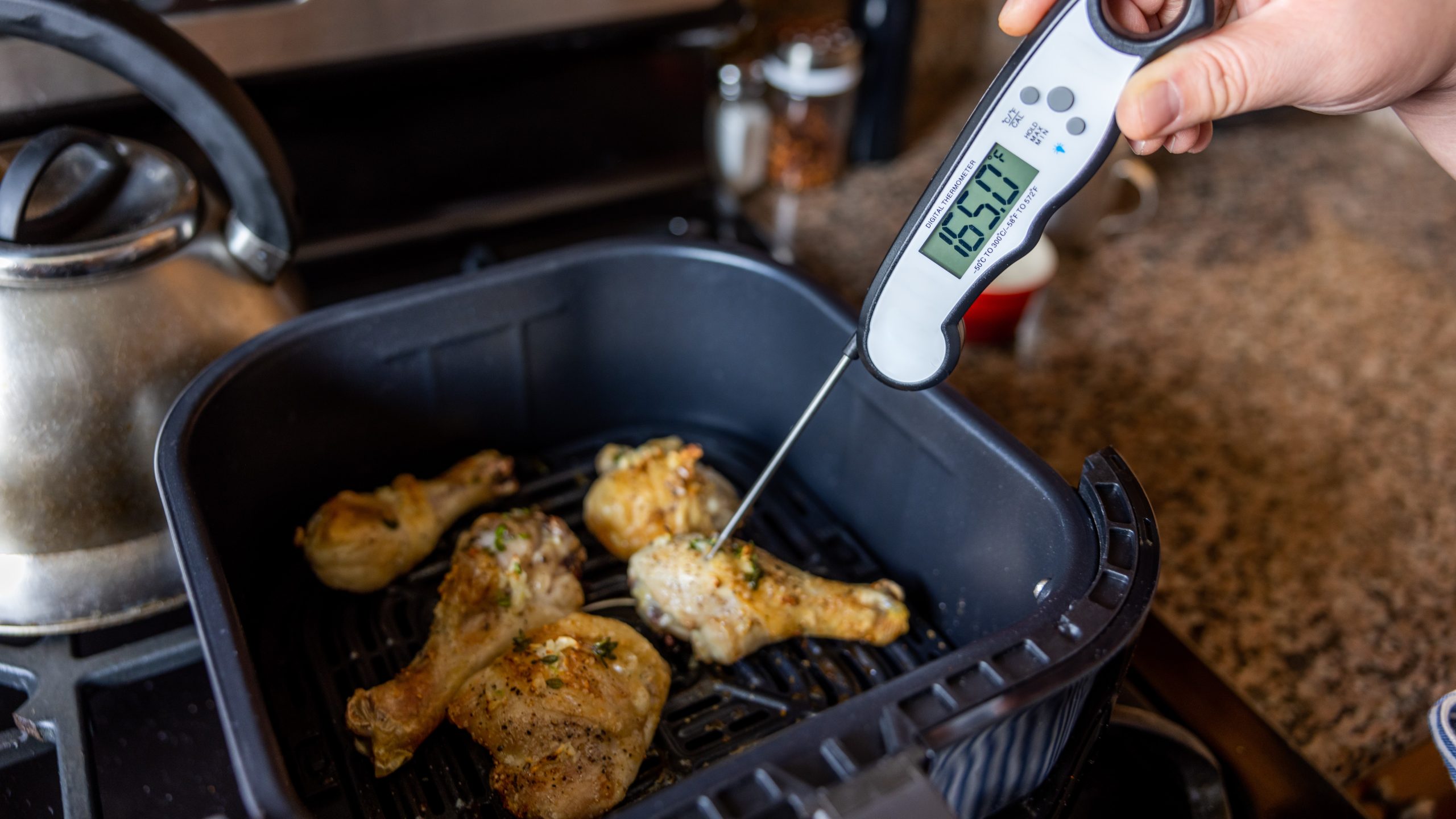 Salmonella - Prevention - Thermometer Person inserting food thermometer into chicken pieces in an air fryer to check temperature. Thermometer reads 165°F.