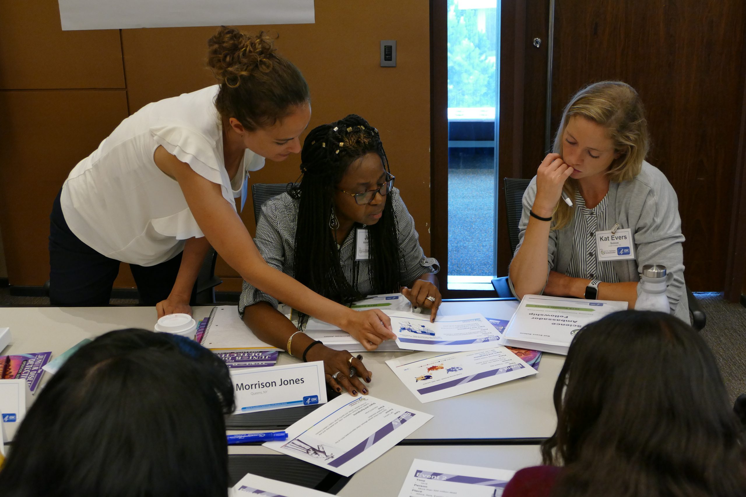 katrina-haynes A dark-haired woman in white shirt works with other teachers. They are at a table covered in educational materials.