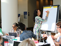 lisa-earehart A dark-haired woman in a colorful dress writes on a giant notepad in a room filled with diverse peers.