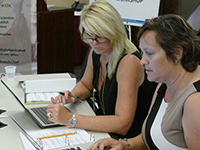 susannah-miller A blonde-haired woman in a black tank top sits at a table in front of a laptop looking at a dark-haired woman's laptop next to her.