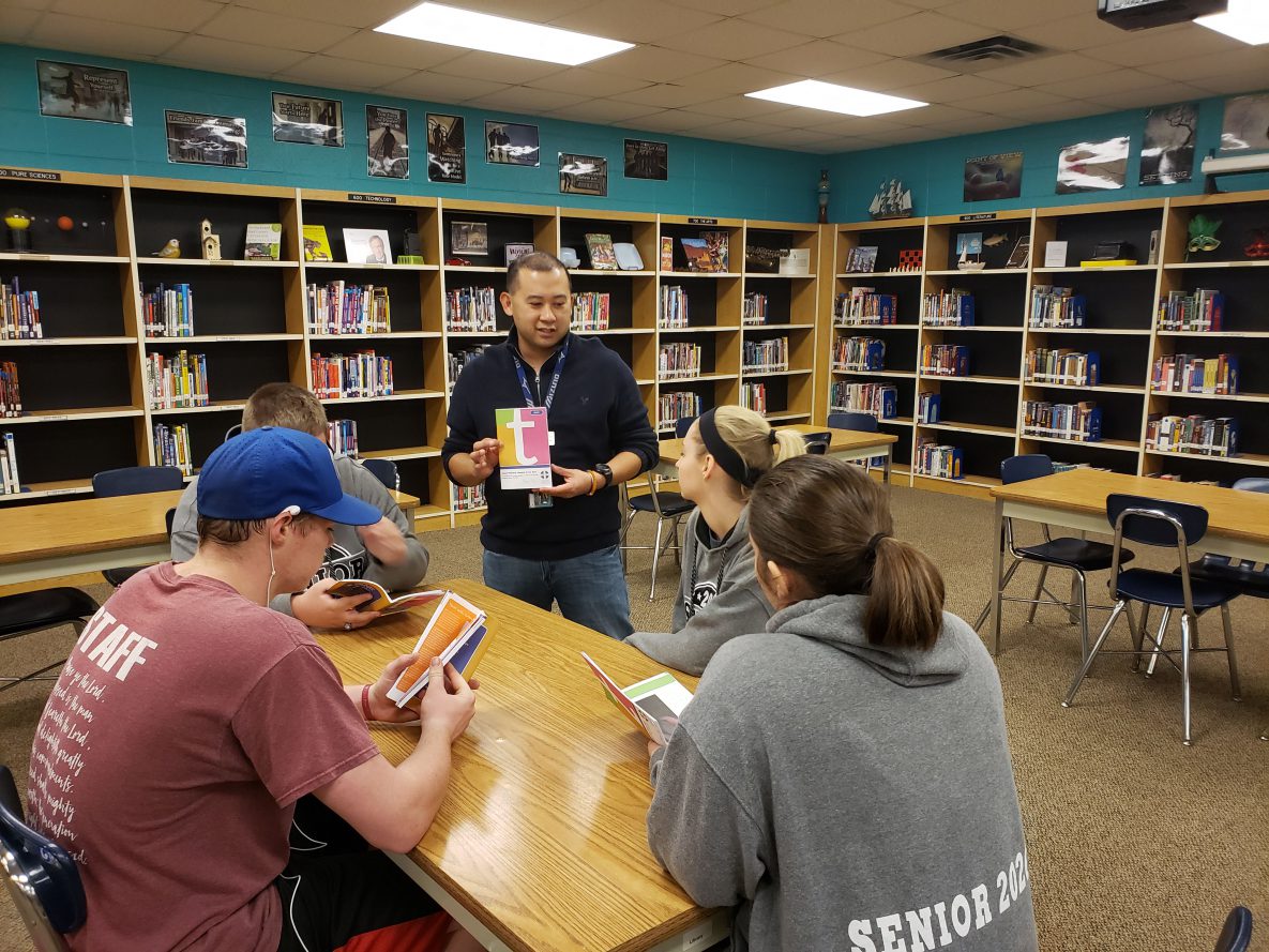Ryan Lacson teacher_students Man standing in a library showing a book to others.