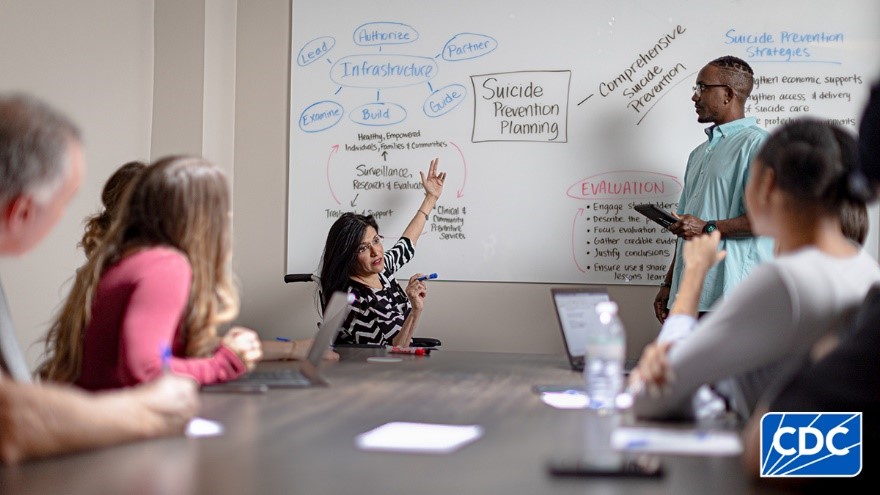 A woman in a meeting pointing to a whiteboard that says suicide prevention planning.