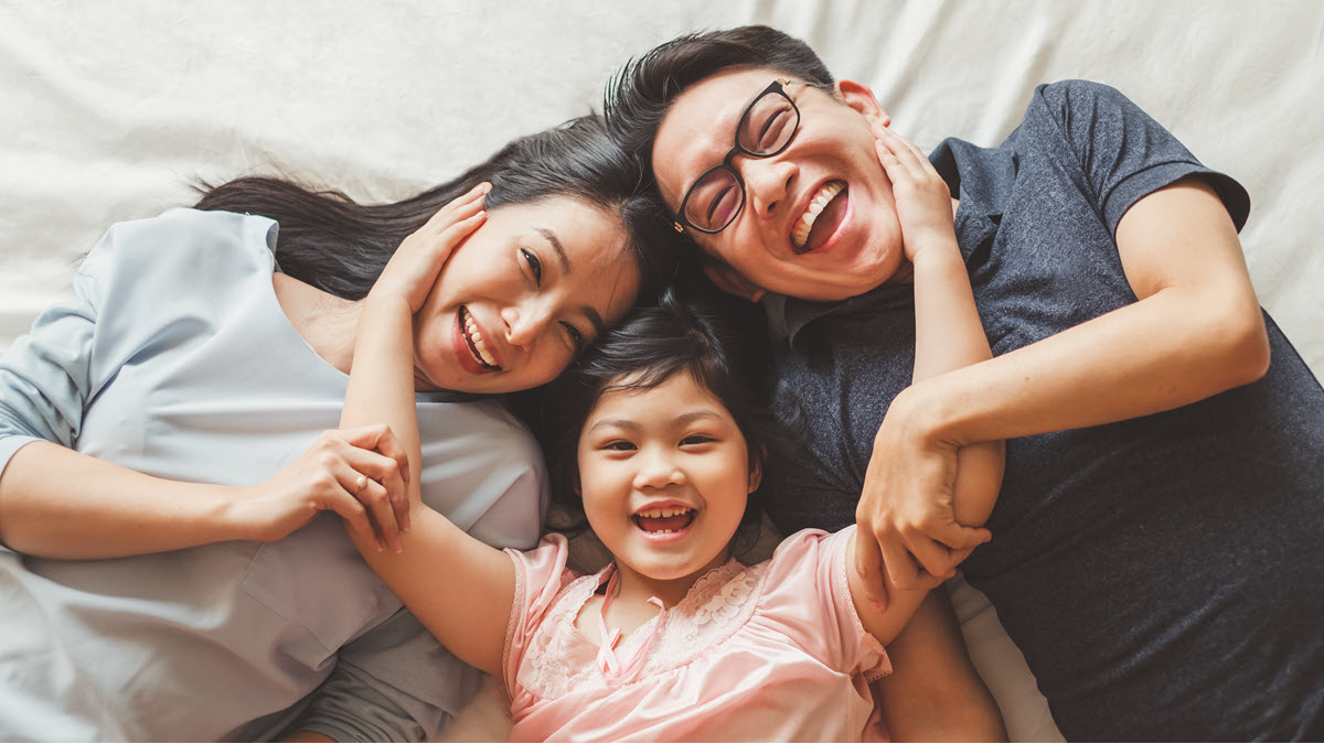 Asian family of three people A happy Asian family laying on a bed laughing with parents and daughter in the middle hugging them