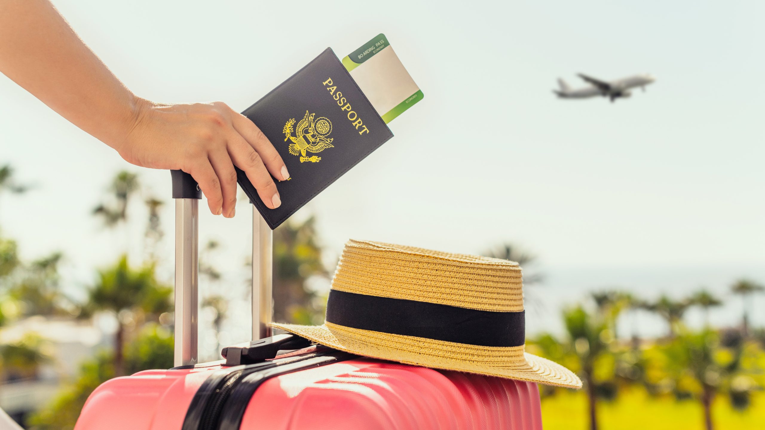 Passport A hand holding a passport and pink suitcase with a hat on top. The person is standing outside watching a plane in the sky.