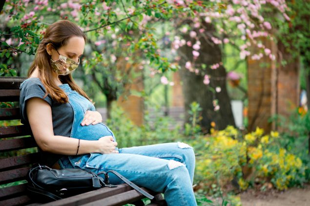 Pregnant vaccination A pregnant woman wearing a cloth facemask sits on a park bench holding her belly.