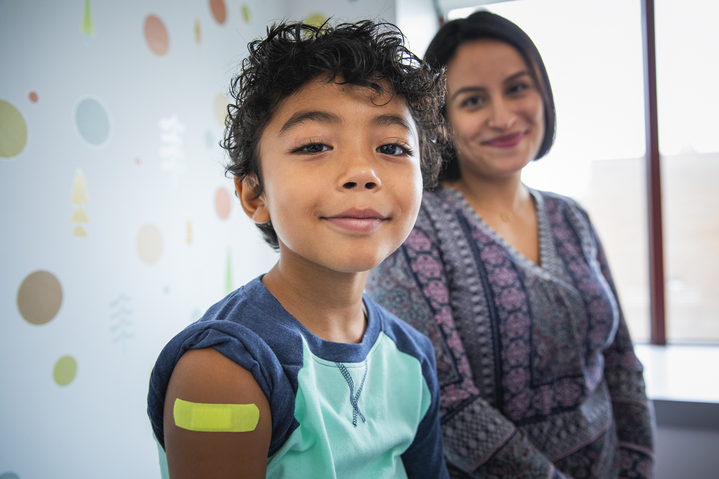 Pediatric patient and Parent Pediatric patient with band-aid on upper arm smiling in medical office with parent smiling in the background.