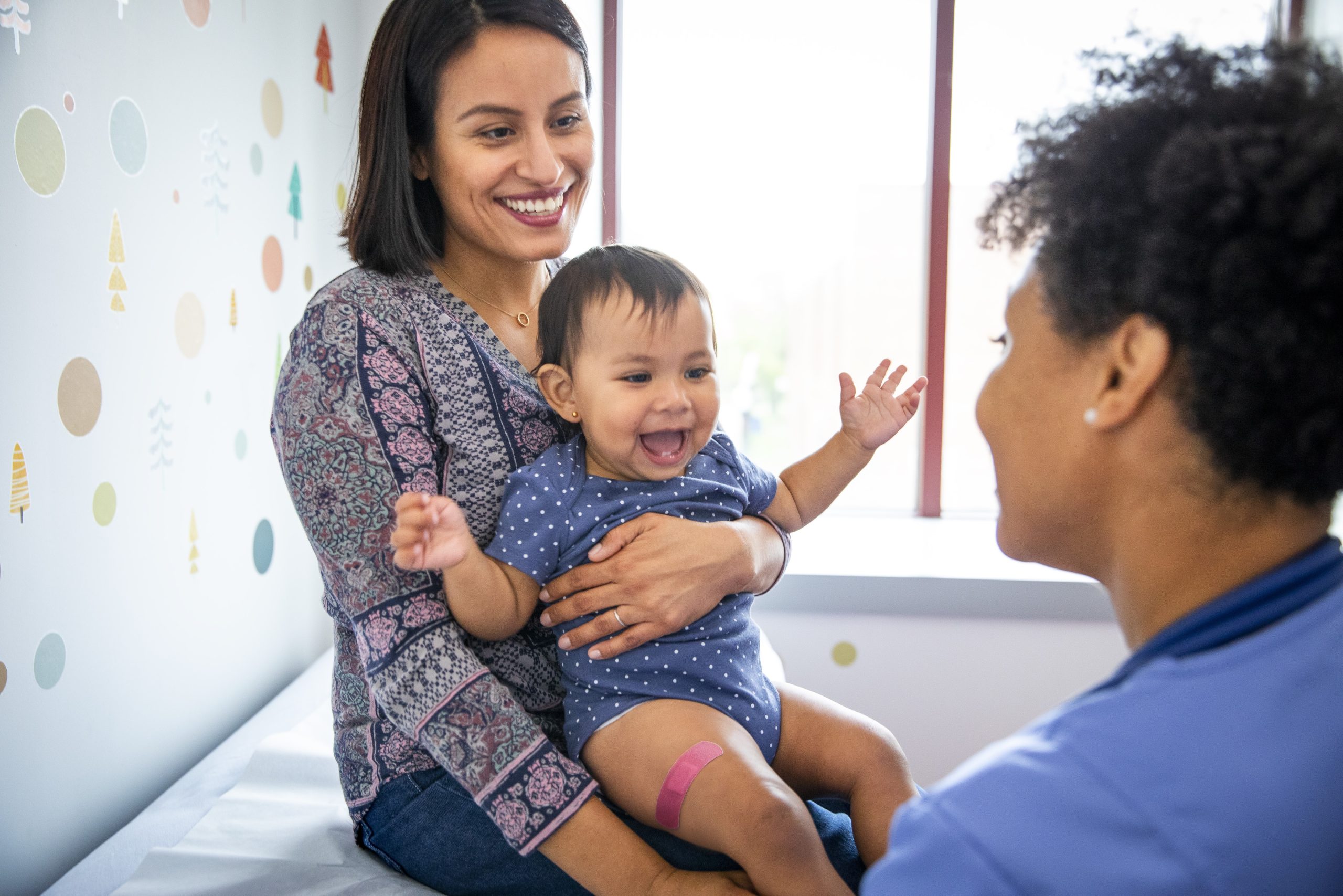 Parent and infant in Medical Office with Healthcare Provider Parent holding infant with a band aid on leg in medical office with healthcare provider post vaccination.