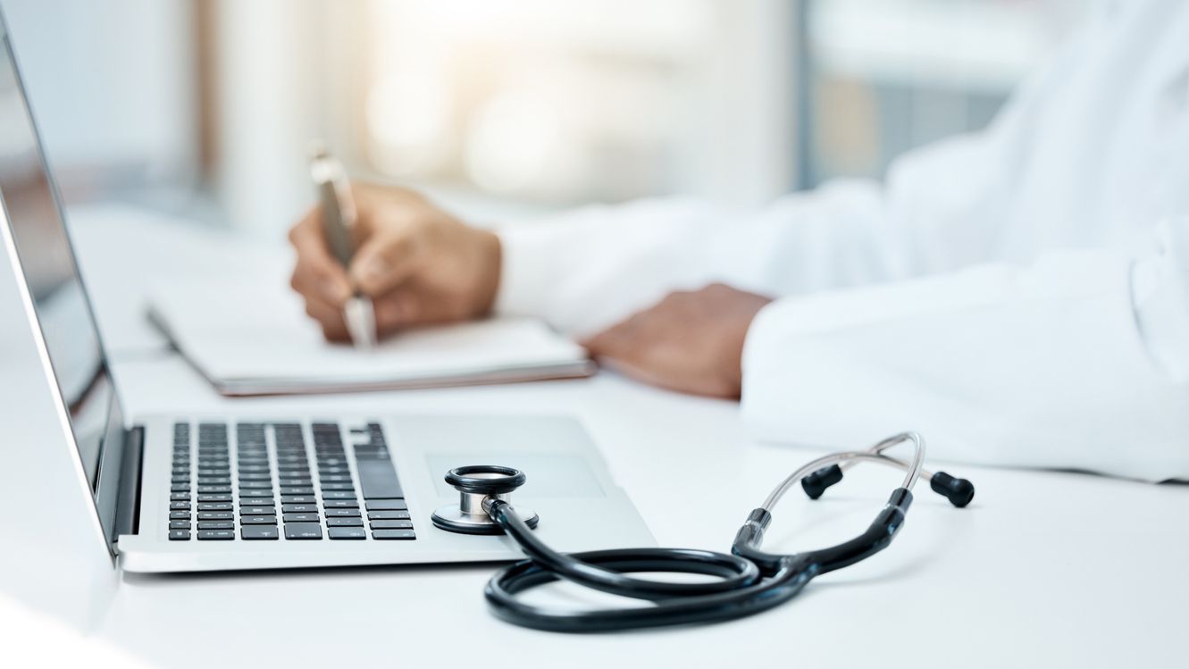 AGE Monitoring Desk with doctor writing on a pad with a computer and stethoscope in the foreground.