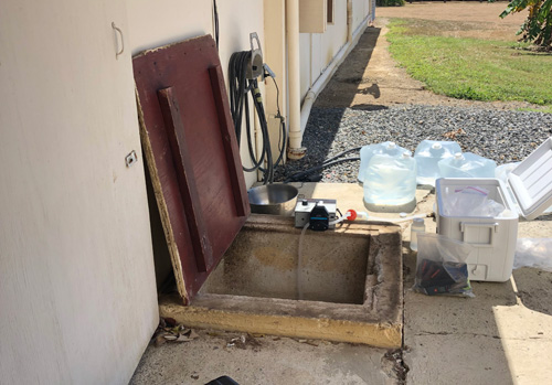 Cisterna subterránea Una cisterna subterránea externa al lado de una edificación. La tapa de madera está abierta y se ve la abertura de concreto cuadrada. Hay una nevera portátil, bolsas de agua y un instrumento para pruebas del agua alrededor de la abertura de la cisterna.