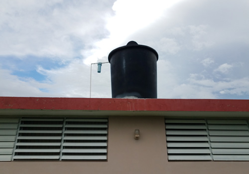 Cisterna en el techo Edificación con un recipiente de almacenamiento de agua grande (cisterna) en el techo. Hay un tubo de plástico entre la cisterna y el techo, con un filtro de agua conectado al tubo.