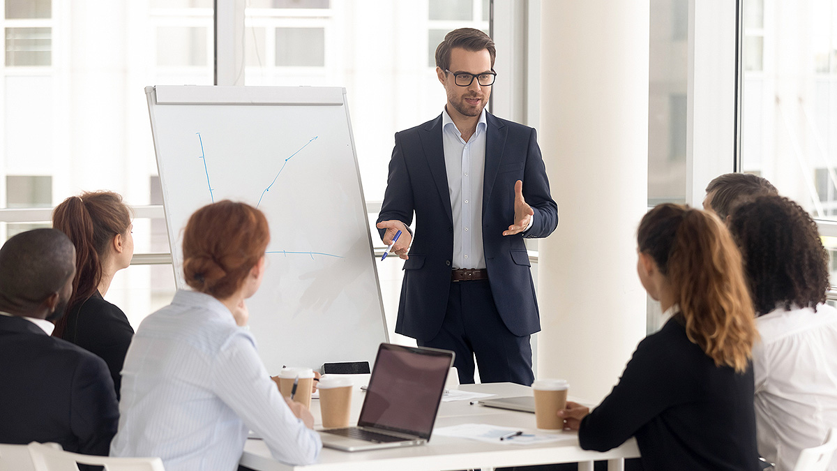Workplace Health Training Chart on Easel Man in suit standing in front of easel with hand-drawn chart in conference room full of employees.