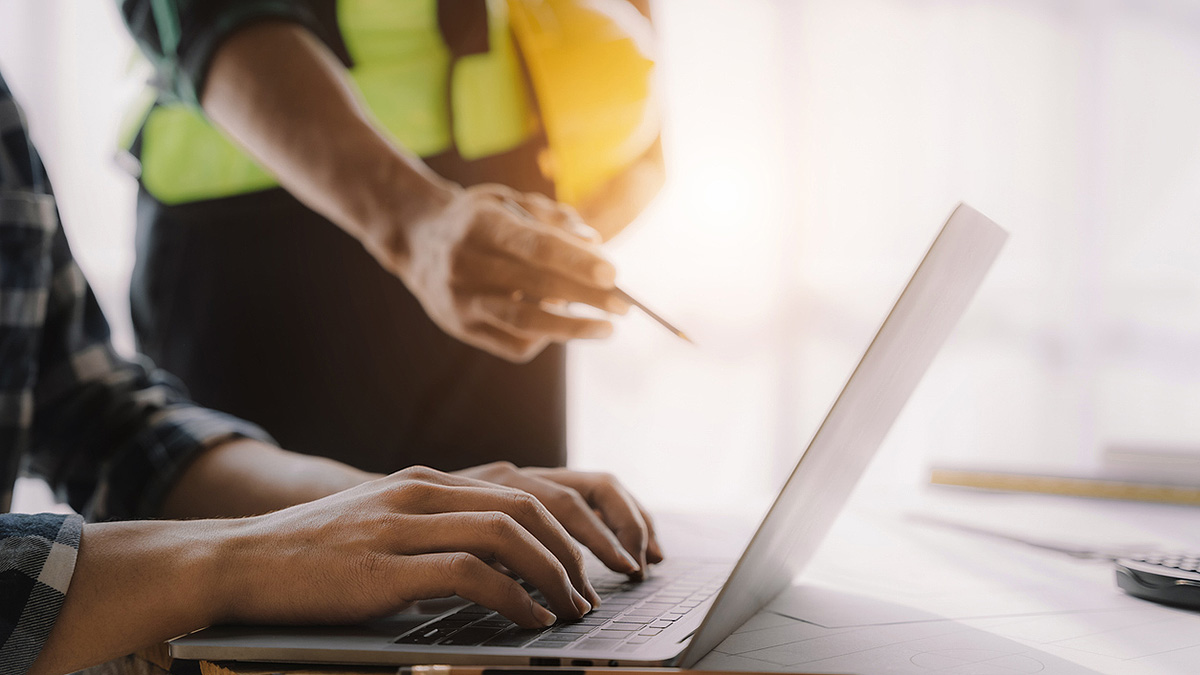 Workplace Health Data on Laptop User imputs data via laptop and worker in construction gear points to screen with pencil.