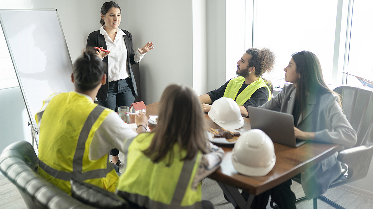 Workplace Health Train the Trainer Businesswoman standing at dry erase board training three workers in hardhat gear and one woman in business attire.