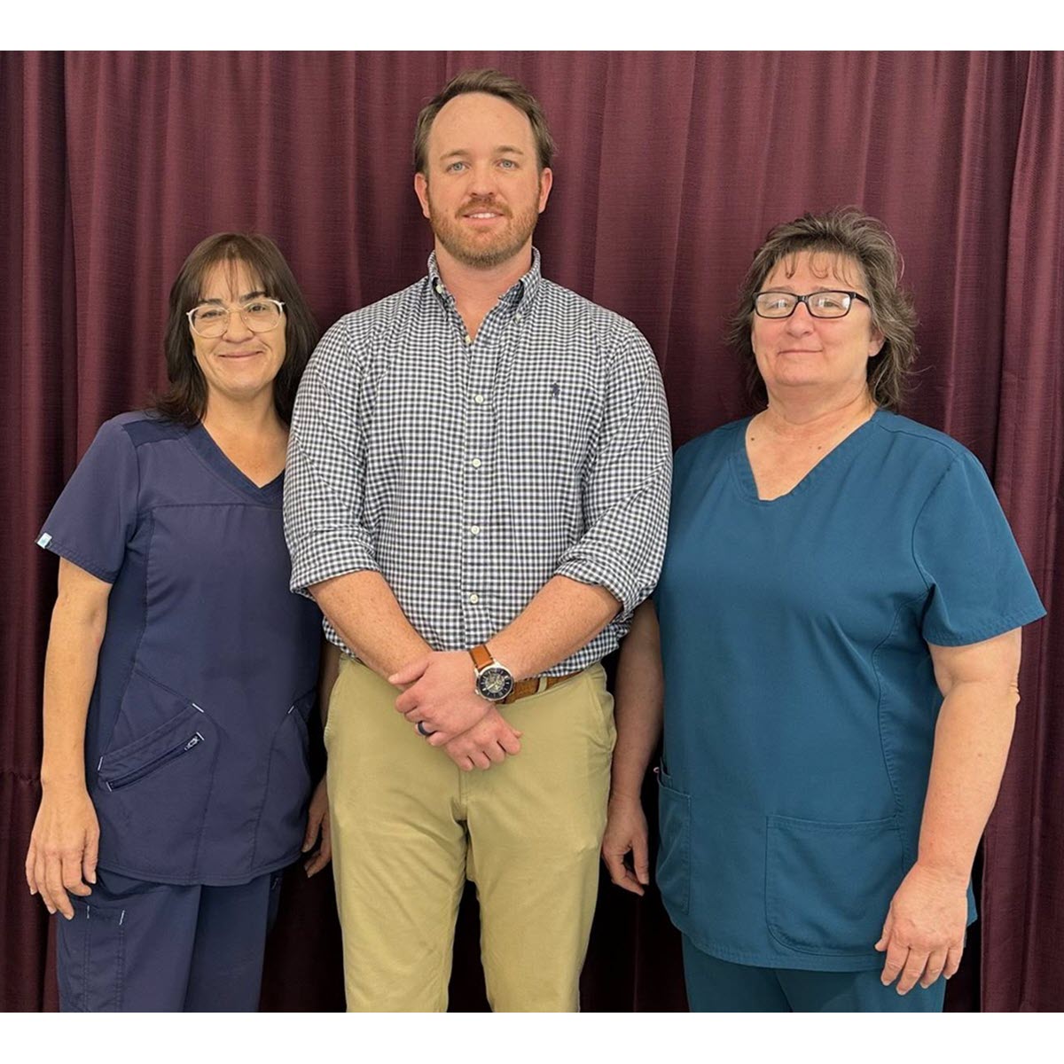 Faith Healthcare 2 women wearing scrubs and a tall man from Faith Healthcare smiling in front of a red curtain