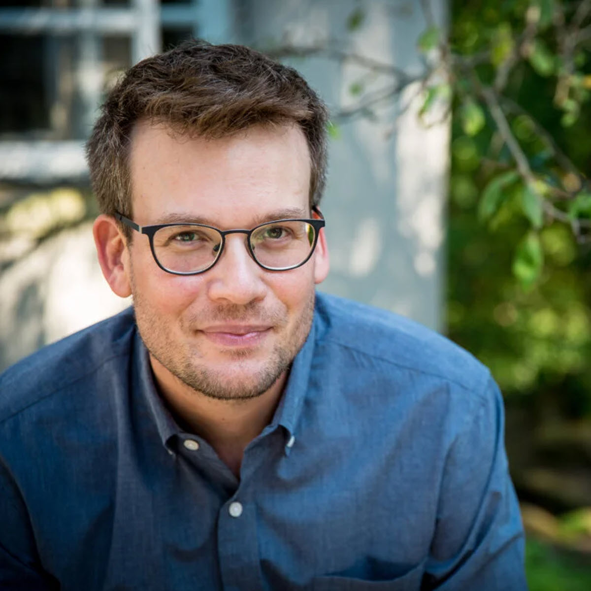John Green John Green smiling outdoors, wearing a blue shirt, glasses, and a beard